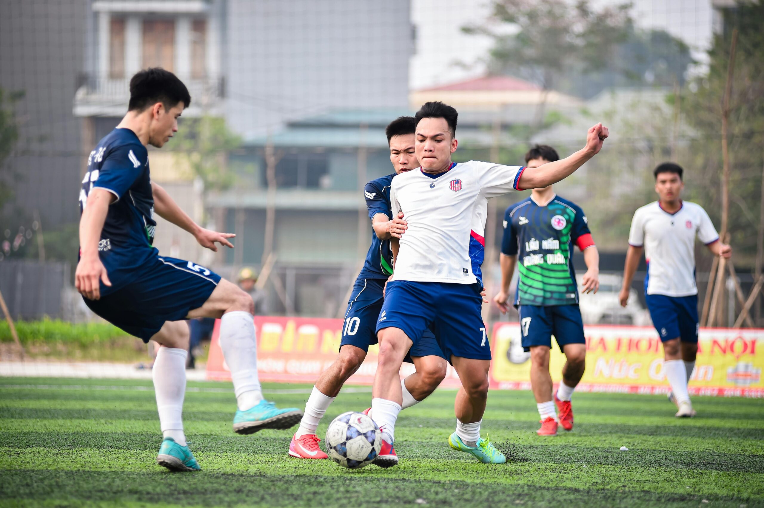 Several young men are playing football, with two engaged in a tackle for the ball and others in the background. They wear football kits in dark blue or white and are playing on an outdoor pitch. This is an example of how ELC Bristol students can play football in the Bristol community. Photo by ANH LÊ: https://www.pexels.com/photo/intense-football-match-action-in-hanoi-35180891/