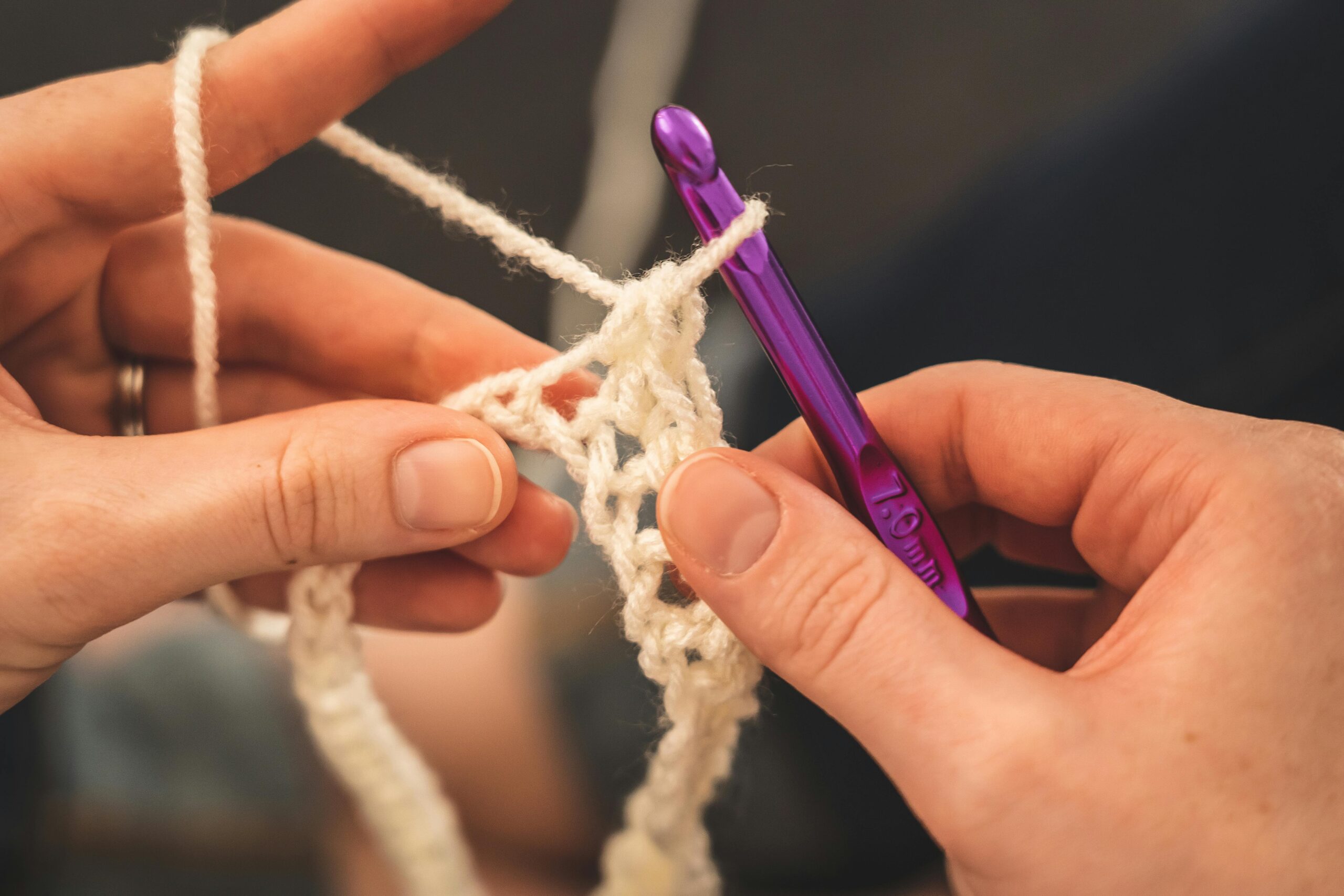 Two hands can be seen close-up as they crochet with white/cream wool.  This is an example of how students can join groups and clubs for knitting and crochet in Bristol.  Photo by Castorly Stock : https://www.pexels.com/photo/person-holding-purple-crochet-hook-and-white-yarn-3945638/