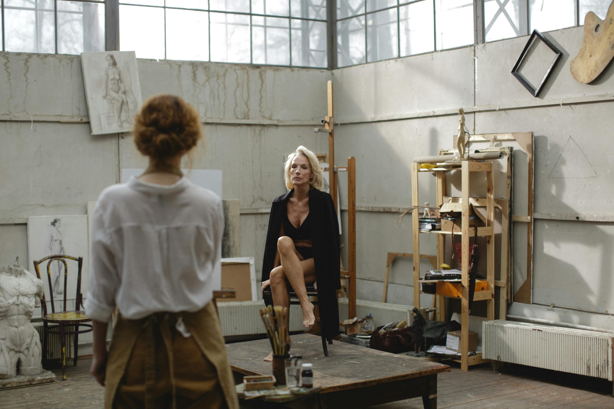 A standing artist draws a seated life model in front of them in a space which looks like an art studio.  Art supplies and life drawings can be seen in the room and on the walls.  The artist has their back to the viewer, but the face of the model can be seen.  This is an example of what to expect from life drawing classes in Bristol.  Photo by cottonbro studio: https://www.pexels.com/photo/artist-drawing-woman-in-black-clothing-seated-on-wooden-chair-3778137/