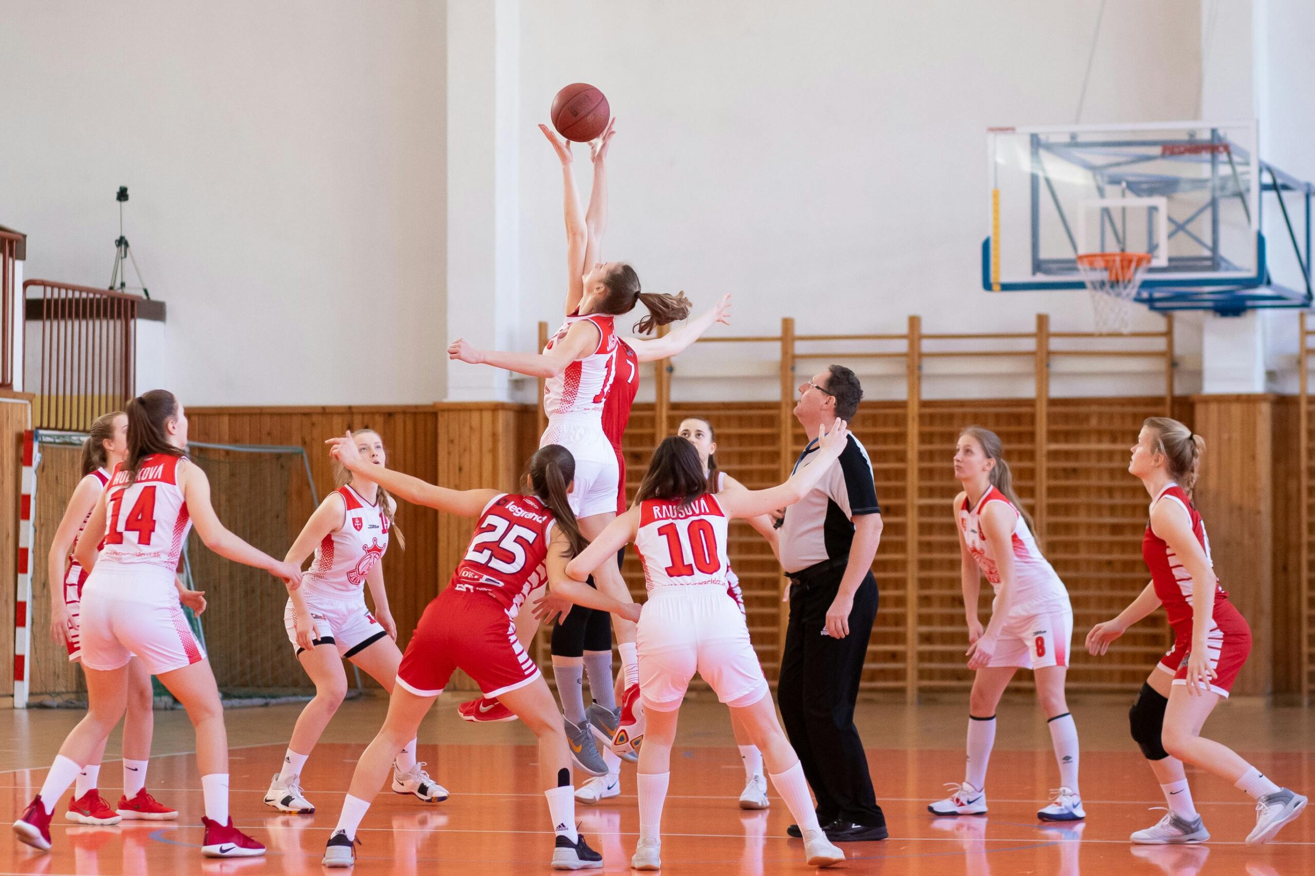 The image shows several members of two basketball teams, playing basketball in an indoor gym.  One player is being propped up by her teammates to try and shoot the ball into the hoop, while other players stand below, ready for the next move.  This is an example of what students can expect if they join a team to play basketball in Bristol.  Photo by IMG_1979 Števonka: https://www.pexels.com/photo/women-playing-basketball-2116469/