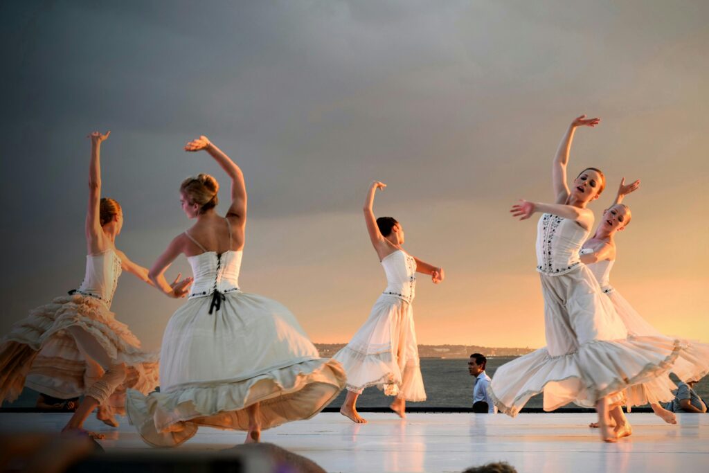 5 women dressed in long white dresses dance in a classical style on a beach over the water. This is just one example of the many opportunities for dancing in Bristol. Photo by Tim Gouw: https://www.pexels.com/photo/5-women-in-white-dress-dancing-under-gray-sky-during-sunset-175658/