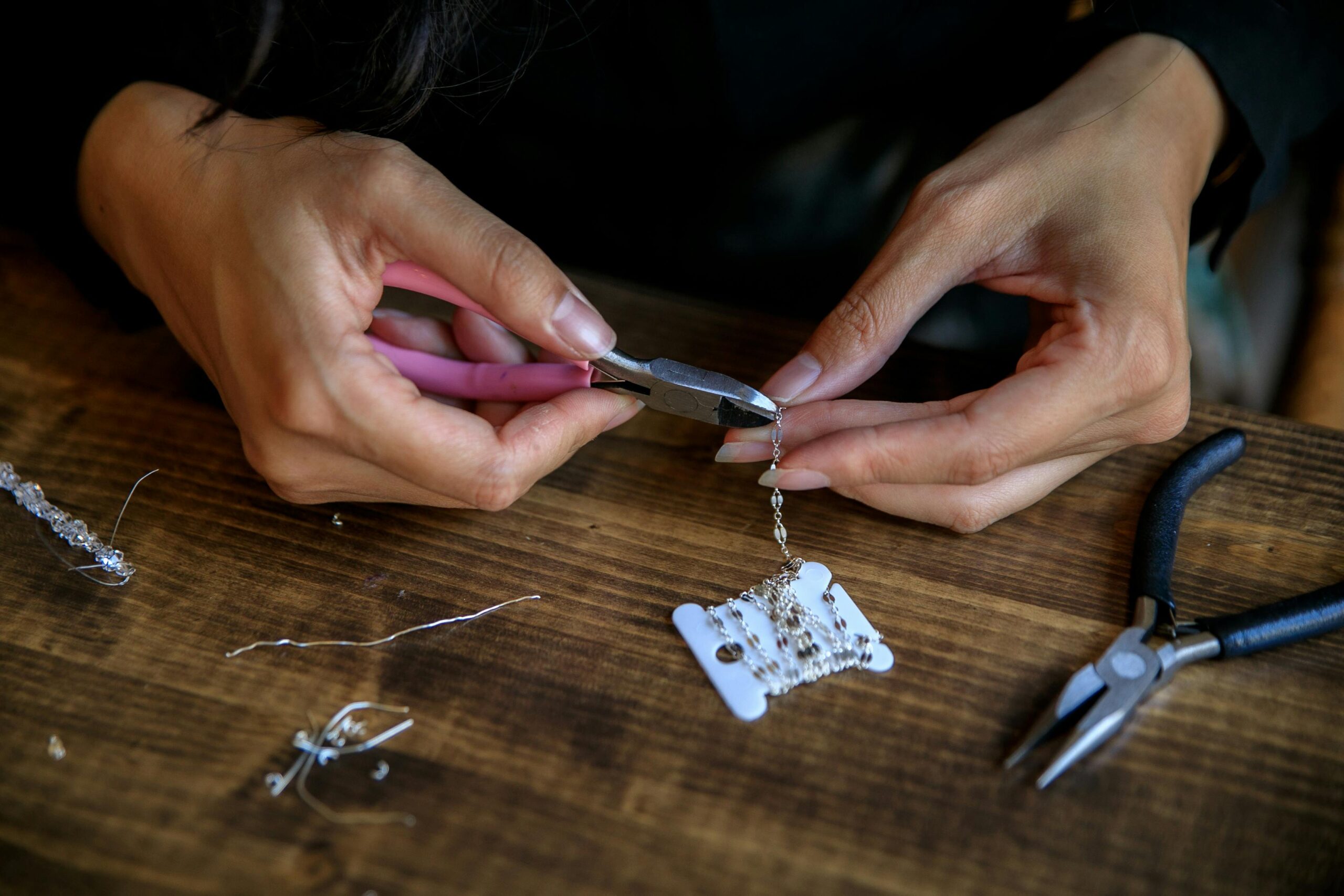 Two hands are seen with some long-nose pliers as they clamp together a jewellery chain, with other jewellery making tools visible on the table.  This is an example of how ELC students can get involved with jewellery making classes in Bristol.  Photo by RDNE Stock project: https://www.pexels.com/photo/using-tools-while-making-handmade-jewellery-10474305/